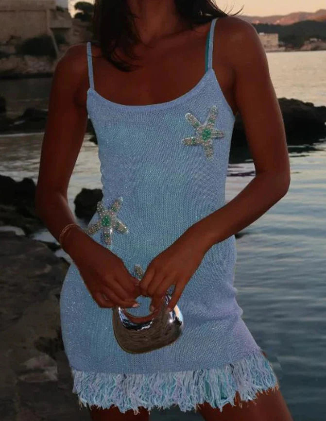Woman wearing a light blue dress with starfish designs by a body of water.