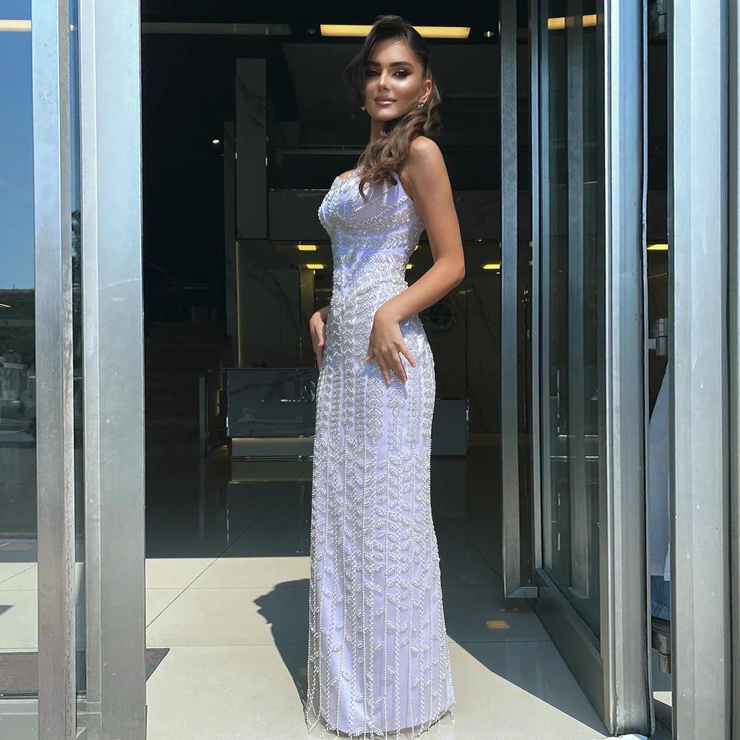 Woman in a white evening gown standing in front of a modern building entrance.