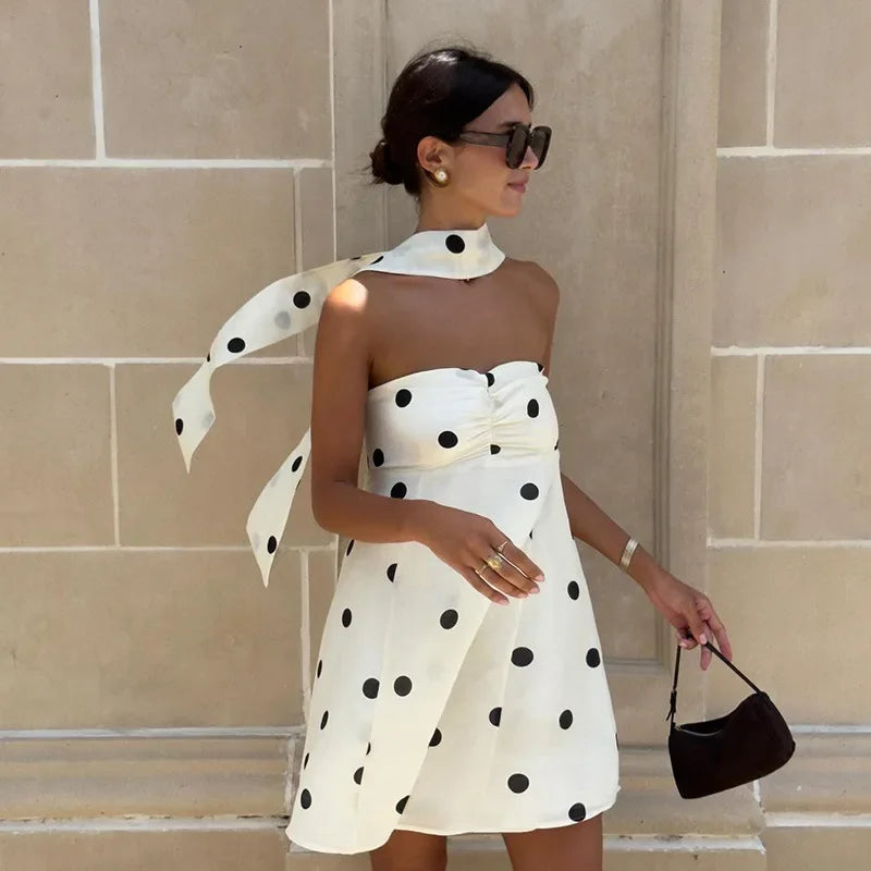 Woman wearing a white polka dot dress with a black handbag against a beige wall.