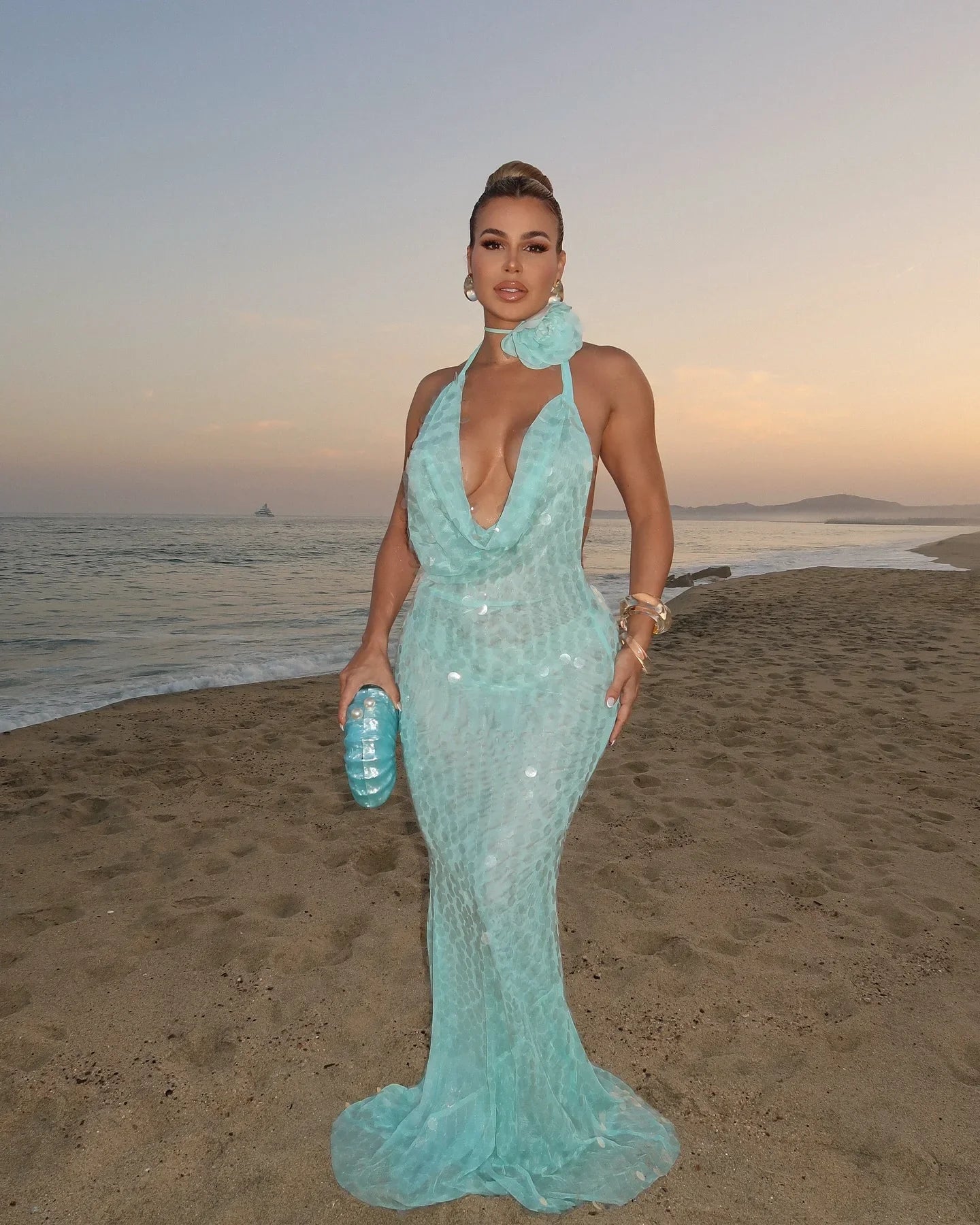 Woman in a light blue evening gown standing on a beach at sunset.