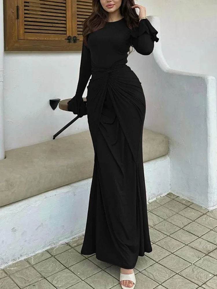 Woman wearing a black long-sleeve dress standing in a room with a wooden cabinet and tiled floor.