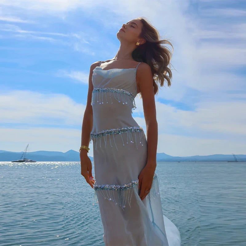 Woman in a white dress with beaded details standing by the water under a blue sky.