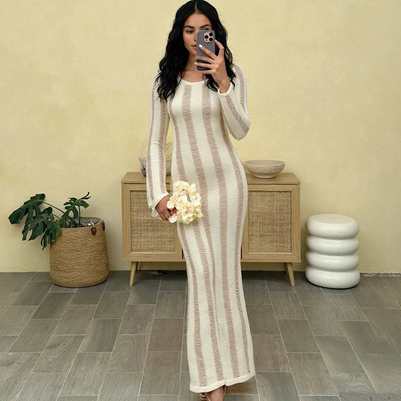 Woman taking a mirror selfie wearing a long, light-colored dress with vertical stripes in a room with a beige wall and wooden cabinet.