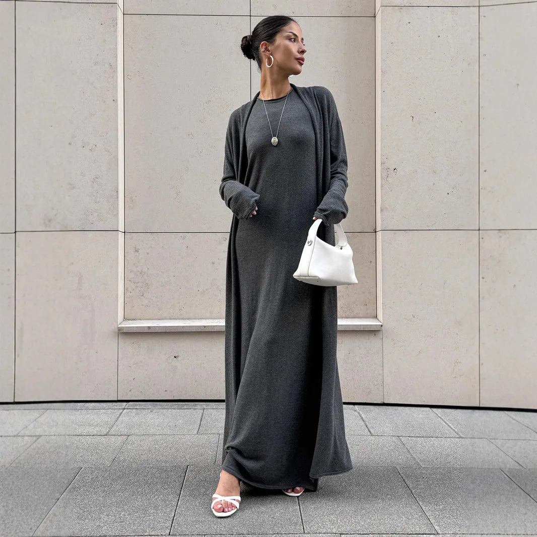 Woman in a long gray dress standing against a light-colored wall.